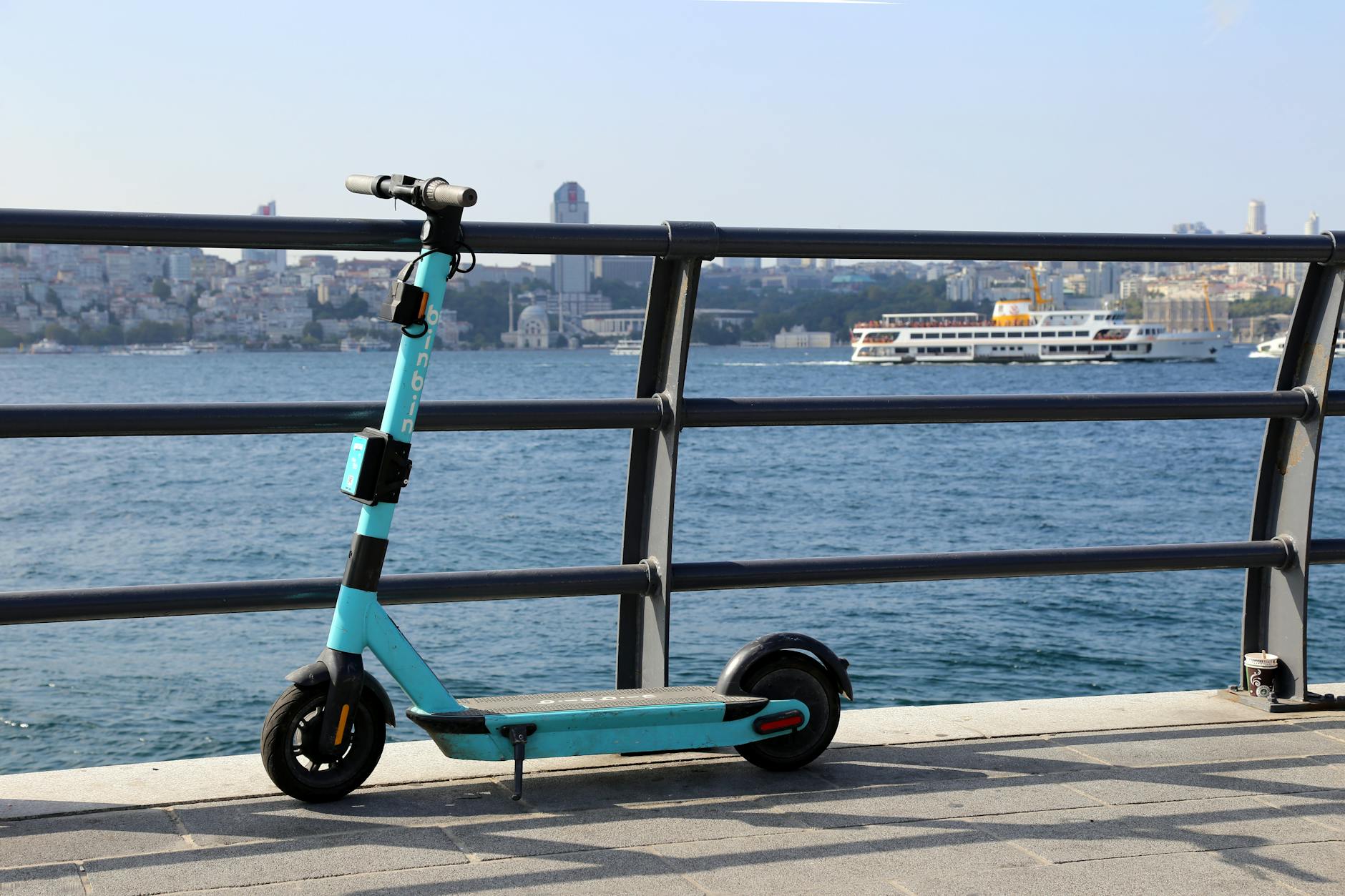 Turquoise electric kick scooter parked by a waterfront with city skyline in background
